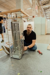 A man with a beard is kneeling on the ground next to a partially assembled metallic structure, possibly a shelf or display rack, inside a spacious industrial or workshop setting. He appears to be working on the assembly with tools nearby, including pliers. The environment is spacious with high ceilings and visible beams.
