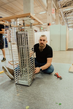 A man with a beard is kneeling on the ground next to a partially assembled metallic structure, possibly a shelf or display rack, inside a spacious industrial or workshop setting. He appears to be working on the assembly with tools nearby, including pliers. The environment is spacious with high ceilings and visible beams.