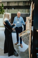 Two people are engaged in conversation in an indoor setting with industrial decor. The woman is wearing a black outfit while the man is dressed in a blue shirt with a name tag. They are standing near a display wall with wooden paneling and a wicker basket filled with stones.