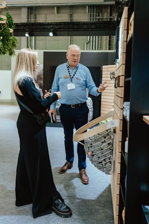 Two people are engaged in conversation in an indoor setting with industrial decor. The woman is wearing a black outfit while the man is dressed in a blue shirt with a name tag. They are standing near a display wall with wooden paneling and a wicker basket filled with stones.