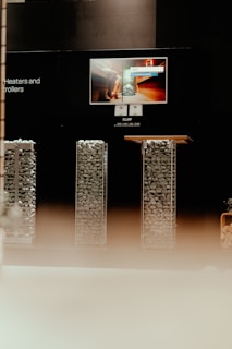 A display setup featuring a modern presentation of heating equipment. At the center, an illuminated screen shows an advertisement for sauna heaters, with a picture of a sauna interior. Below the screen, there are two tall, rectangular structures filled with evenly stacked stones, likely part of the heating equipment being showcased. The environment is dimly lit, creating a focus on the display elements.