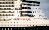 A close-up view of a large cruise ship featuring multiple rows of windows and balconies. The ship is predominantly white with dark accents and has the word 'CUNARD' prominently displayed in red letters on the side.