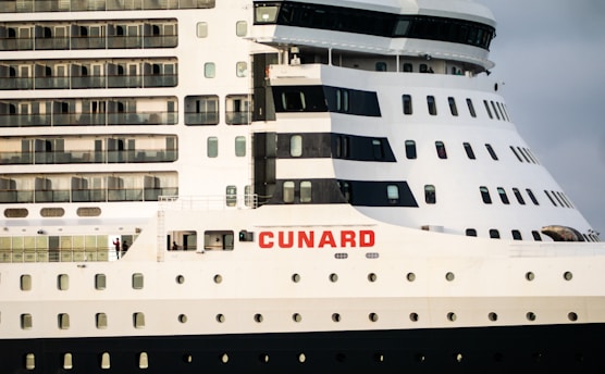 A close-up view of a large cruise ship featuring multiple rows of windows and balconies. The ship is predominantly white with dark accents and has the word 'CUNARD' prominently displayed in red letters on the side.