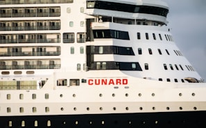 A close-up view of a large cruise ship featuring multiple rows of windows and balconies. The ship is predominantly white with dark accents and has the word 'CUNARD' prominently displayed in red letters on the side.