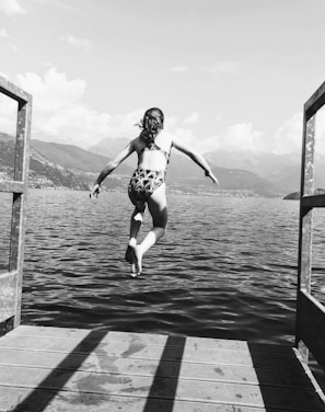 A person is mid-air in a dive over a large body of water, viewed from behind. They are jumping from a wooden platform with railings on either side. Mountains and clouds are visible in the distance under a clear sky.