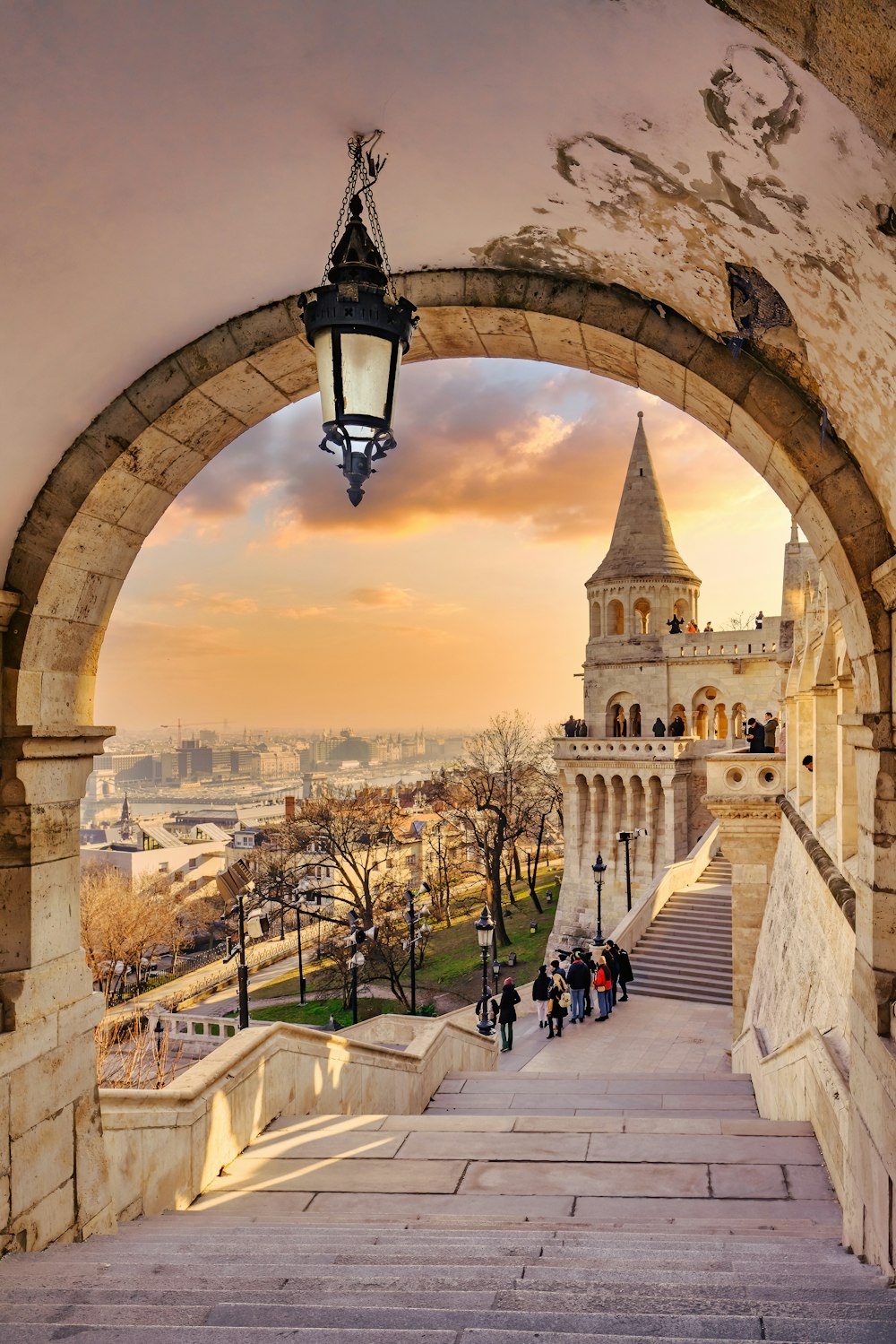 a stone walkway with a lamp post and a large stone arch with a city in the background