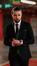A man with dark hair slicked back, wearing a black suit with a white shirt and a black tie. His hands are clasped together, and he stands in an indoor setting with a blurred background featuring some lights and indistinct shapes.