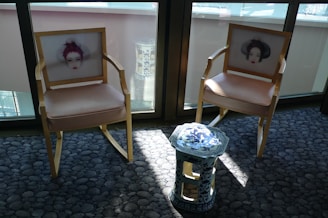 Two wooden chairs with beige cushions feature artistic prints of faces on their backrests. A small, ornate blue and white ceramic stool stands between them. The furniture is placed on a textured grey carpet in a sunlit room, with large windows in the background.