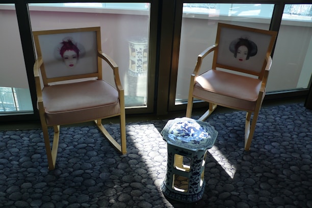 Two wooden chairs with beige cushions feature artistic prints of faces on their backrests. A small, ornate blue and white ceramic stool stands between them. The furniture is placed on a textured grey carpet in a sunlit room, with large windows in the background.
