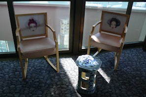 Two wooden chairs with beige cushions feature artistic prints of faces on their backrests. A small, ornate blue and white ceramic stool stands between them. The furniture is placed on a textured grey carpet in a sunlit room, with large windows in the background.