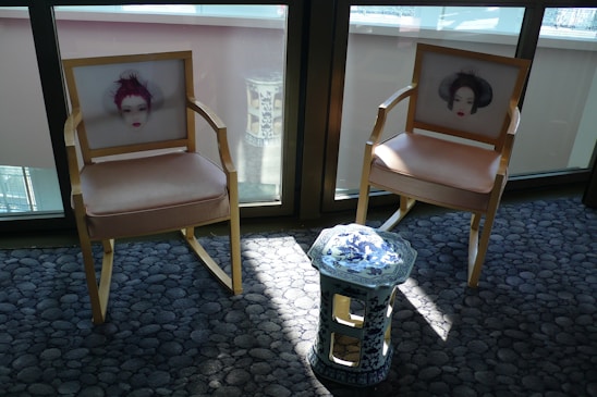 Two wooden chairs with beige cushions feature artistic prints of faces on their backrests. A small, ornate blue and white ceramic stool stands between them. The furniture is placed on a textured grey carpet in a sunlit room, with large windows in the background.