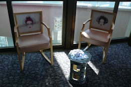 Two wooden chairs with beige cushions feature artistic prints of faces on their backrests. A small, ornate blue and white ceramic stool stands between them. The furniture is placed on a textured grey carpet in a sunlit room, with large windows in the background.