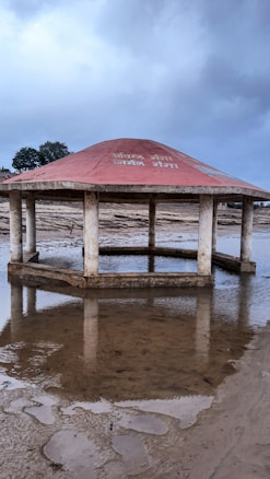 A hexagonal pavilion structure with a red roof stands amidst a muddy and partially waterlogged area. The roof displays text in white. The scene is overcast with a cloudy sky and some trees visible in the background.