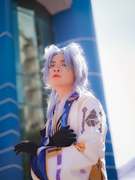 A person is dressed in a detailed costume with long, light-colored hair, animal ears, and decorative clothing. They are wearing black gloves and standing against a backdrop of modern architecture with bright blue and reflective surfaces.
