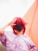 A person with red hair is wearing a face mask and a shirt featuring anime-style illustrations with the word 'Shogun'. The background includes a bright, soft gradient of colors.