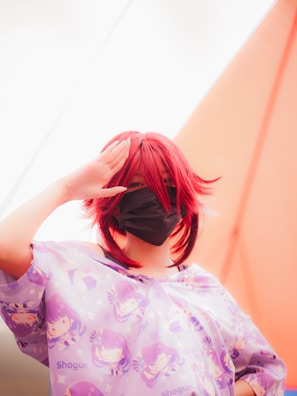 A person with red hair is wearing a face mask and a shirt featuring anime-style illustrations with the word 'Shogun'. The background includes a bright, soft gradient of colors.