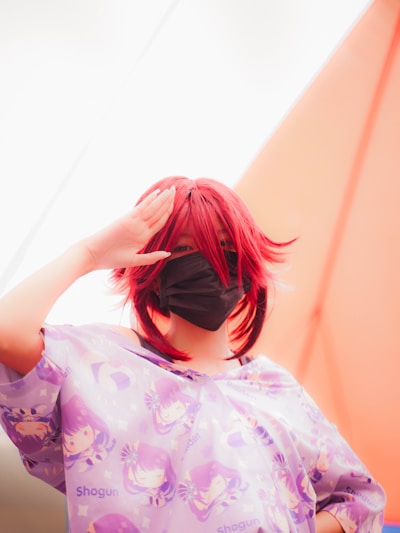 A person with red hair is wearing a face mask and a shirt featuring anime-style illustrations with the word 'Shogun'. The background includes a bright, soft gradient of colors.