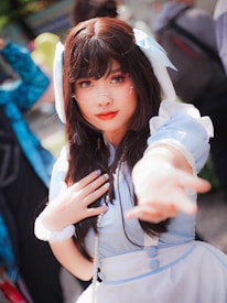 A person with long dark hair is wearing a light blue and white outfit that resembles a maid or anime cosplay costume. They have glasses, bright makeup, and are extending one hand toward the camera. The background shows other people and is blurred, indicating an outdoor gathering or event.