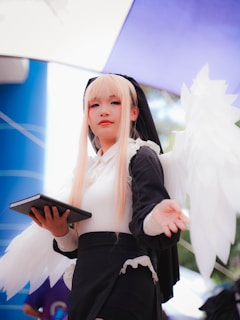 A person dressed in an angelic costume with long blonde hair, holding a tablet in one hand and extending the other hand towards the camera. They have white angel wings and wear a black and white outfit, including a headpiece and skirt. The background includes blue and purple shades that suggest an outdoor setting.