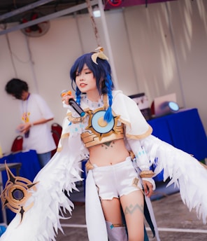 A person is dressed in an elaborate costume with blue braided hair, holding a microphone. The outfit features white, gold, and blue elements with feather-like details. The setting appears to be an indoor or covered area with a purple and white background.