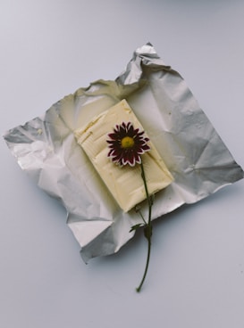 A block of butter rests on an unwrapped piece of foil, topped with a single purple and white flower. The setting is minimalistic with a gentle play of light and shadows.