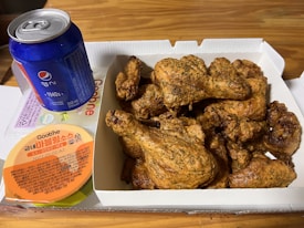 A box of crispy fried chicken pieces is placed on a table, accompanied by a small container of sauce. Next to it, there is a blue Pepsi can. The box has a brand name, 'Goobne,' printed on it.