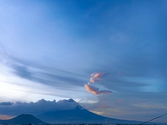 A large mountain is partially covered by clouds, with a clear blue sky above. There is a plume of smoke or cloud rising from the peak, creating a striking pattern. The landscape below is mostly dark, with silhouettes of smaller hills or mountains.