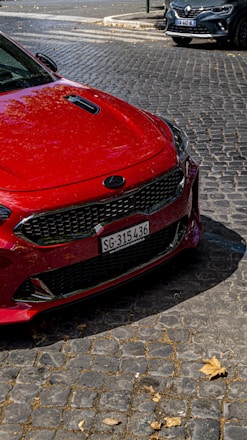 A bright red car with a sleek design is parked on a cobblestone street. The front grille and headlights are prominently visible. Nearby, another car can be seen partially in the background. Fallen leaves are scattered on the cobblestones, hinting at an autumn setting.