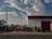 A vintage car and an off-road vehicle are parked on a trailer attached to an old red truck. This setup is situated outside a large industrial building with a red garage door, surrounded by a metal fence. The sky above is partly cloudy with soft, diffuse lighting, casting gentle shadows.