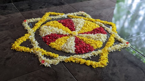 A detailed floral arrangement featuring concentric patterns on a dark tiled surface. The design includes alternating segments of yellow and red flowers, surrounded by layers of white petals. The outermost layer mimics an interconnected star shape with additional yellow petals meticulously arranged.