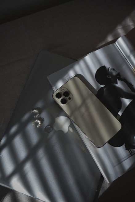 A smartphone with a light-colored back is placed on a laptop, which has a logo visible. Next to them, an open magazine features a black and white photograph of a person in profile. Soft shadows from a window grid fall across the scene, creating an interplay of light and dark patterns. Nearby, a pair of pearl earrings add a touch of elegance.