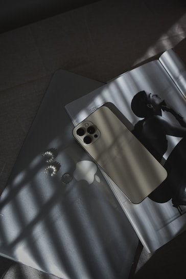 A smartphone with a light-colored back is placed on a laptop, which has a logo visible. Next to them, an open magazine features a black and white photograph of a person in profile. Soft shadows from a window grid fall across the scene, creating an interplay of light and dark patterns. Nearby, a pair of pearl earrings add a touch of elegance.