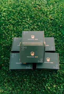 Several black boxes labeled 'Training Box' are stacked on a grassy surface. The top box features illustrations of a kettlebell and a dumbbell.