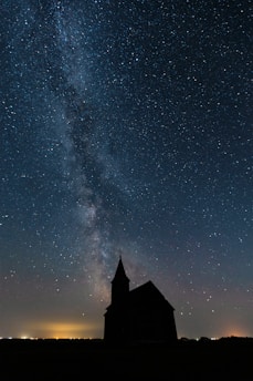 A silhouette of a church is set against a vast, starry night sky with the Milky Way visible. The ground is darkened, and a faint glow from a distant city lights the horizon.