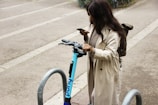 A person is standing near a parked electric scooter, looking at their smartphone. They are wearing a long beige coat and have a backpack. The setting is an urban area, with concrete ground and metal bike racks around.