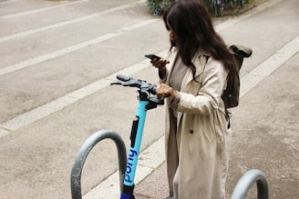A person is standing near a parked electric scooter, looking at their smartphone. They are wearing a long beige coat and have a backpack. The setting is an urban area, with concrete ground and metal bike racks around.