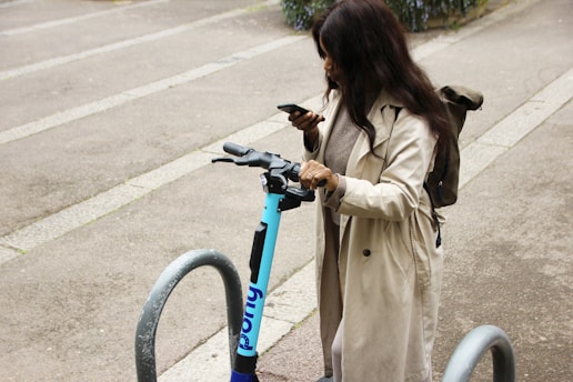 A person is standing near a parked electric scooter, looking at their smartphone. They are wearing a long beige coat and have a backpack. The setting is an urban area, with concrete ground and metal bike racks around.