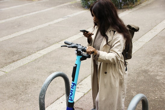 A person is standing near a parked electric scooter, looking at their smartphone. They are wearing a long beige coat and have a backpack. The setting is an urban area, with concrete ground and metal bike racks around.