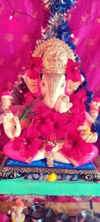 A decorated statue of Lord Ganesha adorned with vivid red flowers. The statue features a detailed golden headdress and is set against a vibrant pink background with additional decorative elements like tinsel. The image conveys a festive and spiritual atmosphere.