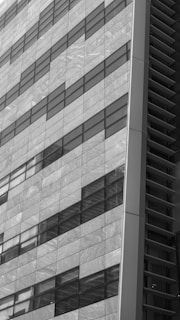 The image features a close-up view of a modern building facade. The surface is composed of large rectangular glass panels arranged in a diagonal pattern. The glass reflects light and the sky, creating varying shades of gray. On the right side, there is a vertical section with metal elements adding contrast and texture to the structure.