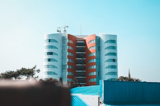 A modern, symmetrical building with a central red vertical section flanked by two white curved structures. The architecture is accentuated by reflective windows that line the facade. In the foreground, there are blue rooftops and trees, creating a contrast with the building. A construction crane is visible on top of the building against a clear blue sky.