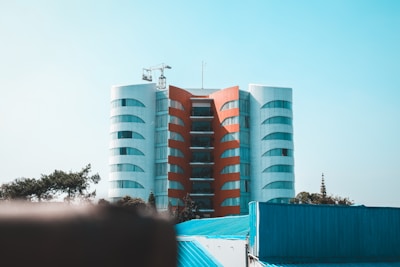 A modern, symmetrical building with a central red vertical section flanked by two white curved structures. The architecture is accentuated by reflective windows that line the facade. In the foreground, there are blue rooftops and trees, creating a contrast with the building. A construction crane is visible on top of the building against a clear blue sky.