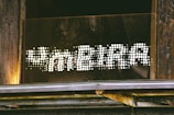 A sign reading 'FAMEIRA' in a dotted, illuminated style is mounted on a wooden and metallic structure. The letters are formed by bright white dots against a dark background, creating a striking contrast. The surrounding area shows weathered wood and some metal elements, suggesting an industrial or rustic setting.