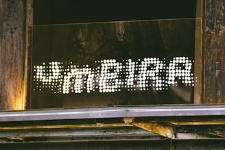 A sign reading 'FAMEIRA' in a dotted, illuminated style is mounted on a wooden and metallic structure. The letters are formed by bright white dots against a dark background, creating a striking contrast. The surrounding area shows weathered wood and some metal elements, suggesting an industrial or rustic setting.