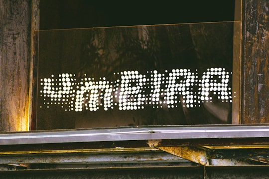 A sign reading 'FAMEIRA' in a dotted, illuminated style is mounted on a wooden and metallic structure. The letters are formed by bright white dots against a dark background, creating a striking contrast. The surrounding area shows weathered wood and some metal elements, suggesting an industrial or rustic setting.