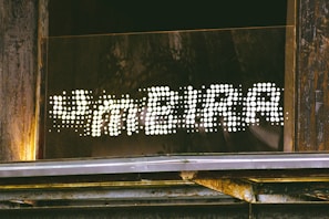 A sign reading 'FAMEIRA' in a dotted, illuminated style is mounted on a wooden and metallic structure. The letters are formed by bright white dots against a dark background, creating a striking contrast. The surrounding area shows weathered wood and some metal elements, suggesting an industrial or rustic setting.