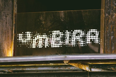 A sign reading 'FAMEIRA' in a dotted, illuminated style is mounted on a wooden and metallic structure. The letters are formed by bright white dots against a dark background, creating a striking contrast. The surrounding area shows weathered wood and some metal elements, suggesting an industrial or rustic setting.