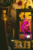 A vibrant street art scene with colorful graffiti featuring a stylized character. In the foreground, a bright pink neon outline of a person stands out, adding contrast to the mural's detailed design. The image includes reflections and lights, creating an urban atmosphere.