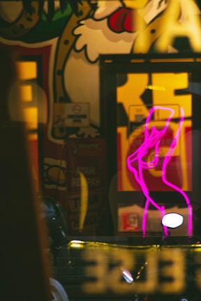A vibrant street art scene with colorful graffiti featuring a stylized character. In the foreground, a bright pink neon outline of a person stands out, adding contrast to the mural's detailed design. The image includes reflections and lights, creating an urban atmosphere.