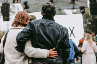A couple standing closely together, with the woman's arm around the man's back. They appear to be attending an outdoor event, visible by a partially blurred crowd and stage setup in the background. The woman is wearing a light-colored coat, while the man is dressed in a dark denim jacket.